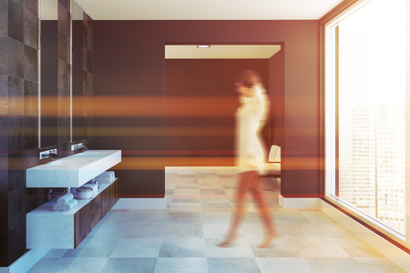 Side view of a white and wooden twin sink vanity unit attached to a black tile bathroom wall with two vertical mirrors hanging above it. A woman walking. 3d rendering toned image blurredの写真素材