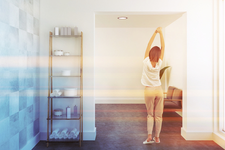 Woman in a bathroom interior with white walls, a concrete floor, shelves with towels and a sofa in the background. 3d rendering mock up toned imageの写真素材