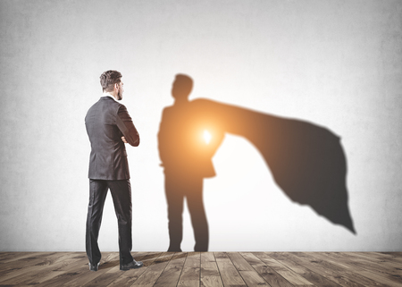 Confident businessman with his hands in pockets looking at his shadow with a cape in a concrete wall room. Concept of leadership and career.の写真素材