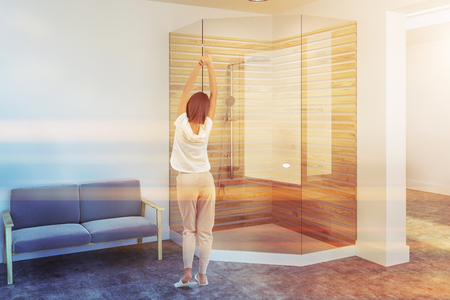 Woman standing near a glass wall shower stall in a white bathroom interior with a concrete floor. A blue sofa in the corner. 3d rendering toned imageの写真素材