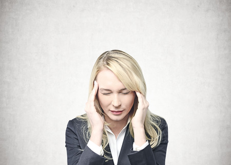 Young caucasian businesswoman with long blond hair wearing a suit having a severe headache. A concrete wall background. Concept of stress and overwork in business. Mock upの写真素材