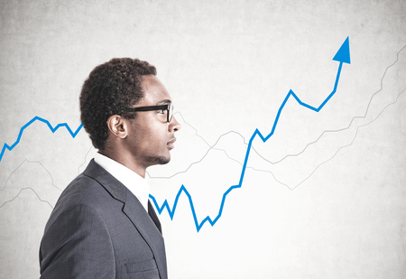 Side view of a young handsome African American businessman wearing glasses and a gray suit. A concrete wall background with a blue growing graph on it.の写真素材