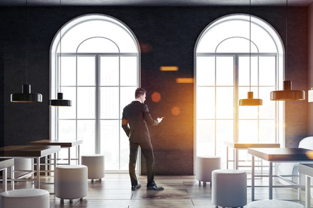 Businessman in a comfortable cafe interior with gray walls, arched windows, square wooden tables and white round chairs. 3d rendering mock up toned imageの写真素材