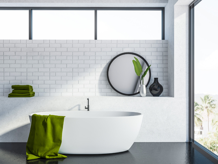 Modern bathroom interior with white brick walls, a gray floor, a white bathtub with a green towel hanging on it and a round mirror. 3d rendering mock upの写真素材