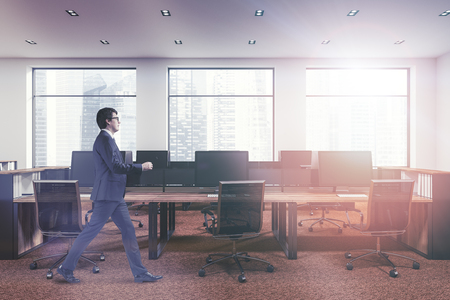 Young businessman walking in open space office of a modern company with white walls, panoramic windows, brown carpet and rows of wooden computer tables. 3d rendering mock up toned imageの写真素材