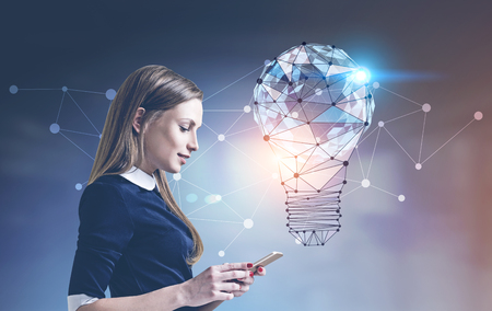 Side view of a smiling calm businesswoman looking at her smartphone screen. A blurred background with a glowing light bulb and a network interface. Toned image double exposureの写真素材