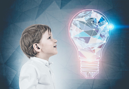 Side view of adorable little caucasian boy with blond hair wearing white shirt and dark blue jeans and looking upwards. Colorful polygonal light bulb background. Creative thining concept toned imageの写真素材