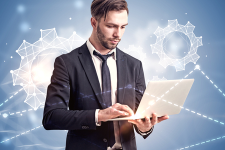 Serious young businessman holding a laptop standing over immersive interface with cogwheels and graphs over blue. Toned image double exposure mock upの写真素材