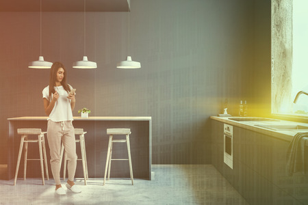 Young woman standing in interior of gray kitchen with concrete floor, big window and grey and wooden countertops. Bar with stools. Toned image double exposureの写真素材