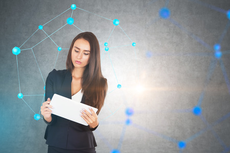Serious european businesswoman in suit looking at tablet computer. Concrete wall and network...