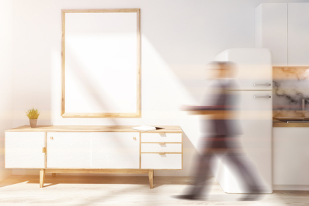 Man walking in kitchen interior with wooden floor, white bridge and countertops, white set of drawers and vertical mock up poster frame. Toned image mock up blurredの写真素材