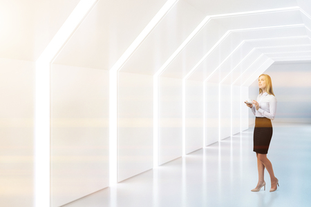 Blonde businesswoman in futuristic empty white hexagonal shaped corridor with lighted ceiling and walls. Side view. Concept of creativity in architecture and hi tech. Toned imageの写真素材