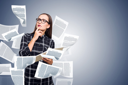 Pensive and attractive young author in glasses and checkered dress standing with copybook in hands and paper flying around over blue background. Creative writing concept. Mock up double exposureの写真素材