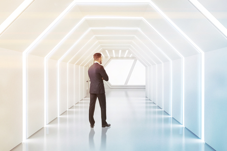 Man in suit thinking in futuristic white hexagonal shaped corridor with lighted ceiling and walls. Side view. Concept of creativity in architecture and hi tech. Toned imageの写真素材