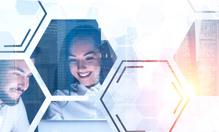 Cheerful young businesswoman and her bearded colleague working together with laptop. Cityscape background. Hexagonal pattern foreground. Toned image double exposureの写真素材