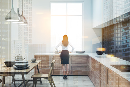Woman in kitchen with white and gray walls, wooden floor, and wooden and white countertops with built in appliances and dishes on them. Wooden talbe with chairs. Toned image double exposureの写真素材