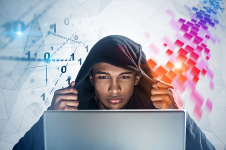 African American hacker looking at his laptop sitting near geometric pattern wall with digital...