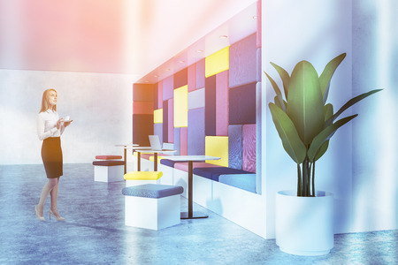 Woman in modern cafe interior with white walls, concrete floor, yellow and red sofa, row of white square tables with red, blue and yellow chairs and a plant in white pot. Toned imageの写真素材