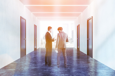 Businessmen talking in interior of office corridor with white walls, closed black doors, concrete floor and large window. Toned imageの写真素材