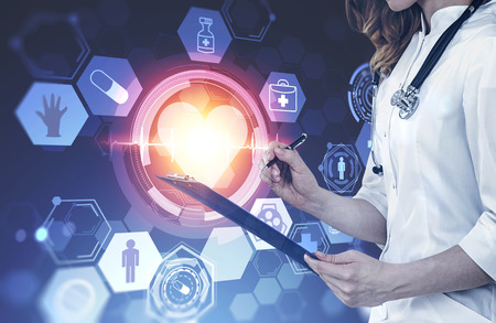 Unrecognizable woman doctor standing over dark blue background next to immersive medical interface with heart in hud. Toned imageの写真素材