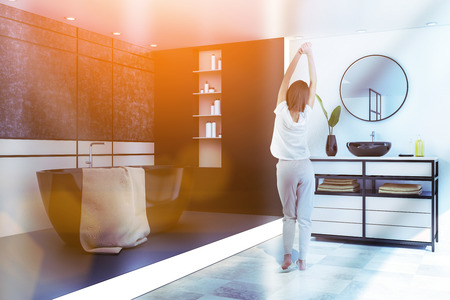Woman in luxury bathroom with beige walls, tiled floor, black bathtub with brown towel on it and sink standing on beige countertop. Toned imageの写真素材