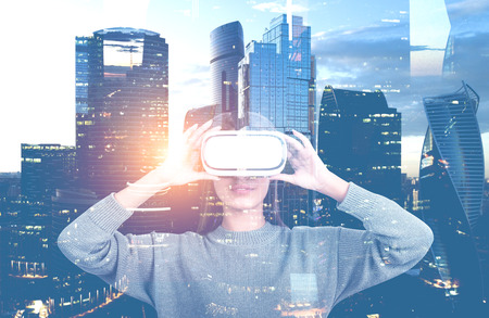 Attractive young woman wearing sweater and vr glasses with double exposure of modern cityscape. Hi...