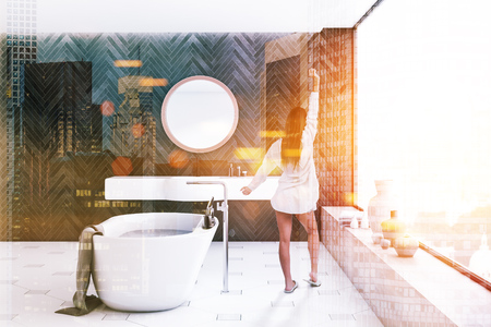 Woman in interior of modern bathroom with black wooden walls, tiled floor, panoramic window, white bathtub and white sink with round mirror above it. Toned image double exposureの写真素材