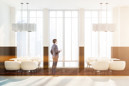 Man in interior of modern cafe with white and brown walls, wooden floor, loft windows and wooden tables with white armchairs. Toned imageの写真素材