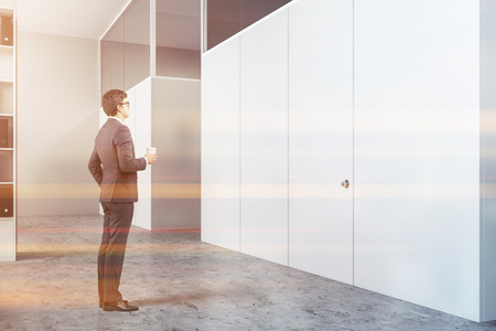 Businessman in interior of modern office with white walls, concrete floor, white bookcase with folders and white doors leading to smaller rooms. Toned imageの写真素材