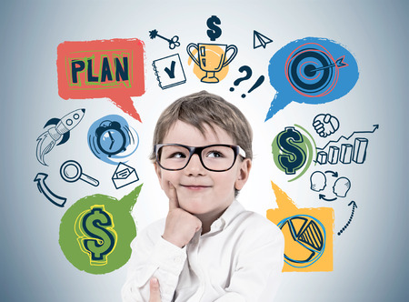 Smiling thinking little boy wearing glasses and white shirt standing near gray wall with colorful business plan sketch drawn on it. Concept of future of businessの写真素材
