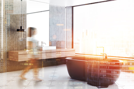 Woman in interior of modern bathroom with concrete walls, tiled floor, panoramic window, gray bathtub and white sink with vertical mirror above it. Toned image double exposure blurの写真素材