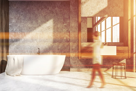 Woman in interior of modern bathroom with concrete and wooden walls, white floor, white bathtub and sink standing on wooden countertop. Toned image blurの写真素材