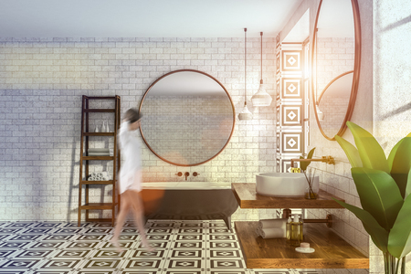 Woman in interior of modern bathroom with brick walls, tiled floor, black bathtub with round mirror and white round sink on wooden shelf. Shelves for towels to the left. Toned image blurの写真素材