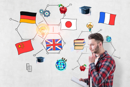 Pensive young man wearing checkered shirt and holding copybook standing near concrete wall with colorful international education sketchの写真素材