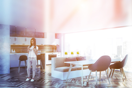 Woman in interior of panoramic kitchen with concrete walls, wooden floor, white countertops, white and wooden bar and table with beige sofa and gray chairs. Toned imageの写真素材