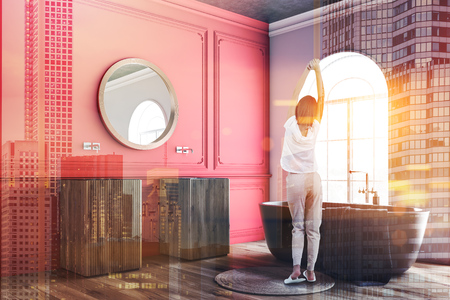 Woman in corner of white and red bathroom with wooden floor, arched windows, wooden double sink with round mirror above it and gray bathtub with towel on it. Toned image double exposureの写真素材