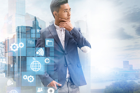 Thoughtful Asian businessman standing over modern cityscape background with double exposure of business icons. Toned imageの写真素材