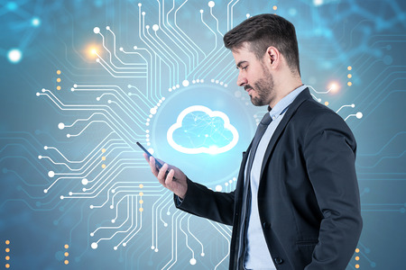 Side view of bearded young businessman looking at his smartphone standing over cloud computer...