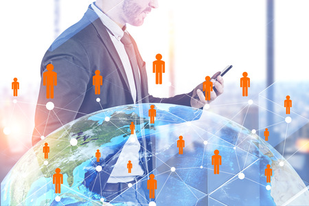 Side view of unrecognizable businessman looking at smartphone standing in office with double exposure of global people network hologram.の写真素材
