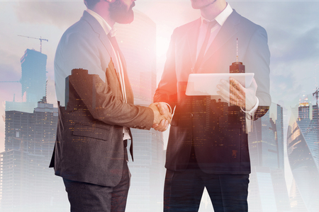 Two unrecognizable businessmen shaking hands. One of them is holding a tablet. Double exposure of night cityscape. Toned imageの写真素材