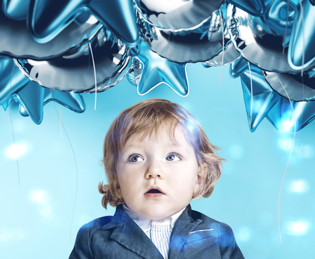 Adorable astonished little boy with blond hair in suit looking at blue balloons standing over light...