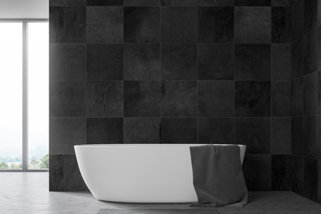 Minimalistic bathroom interior with black tile walls, concrete floor, large window with mountain view and white bathtub with gray towel on it. 3d renderingの写真素材