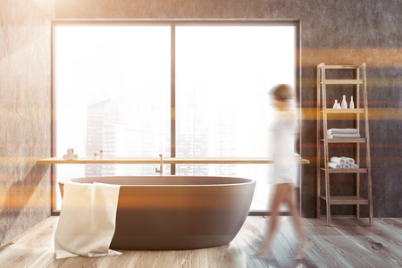 Woman in interior of stylish bathroom with gray walls, wooden floor, gray bathtub with white towel on it and wooden shelves with towels and creams. Toned image blurの写真素材