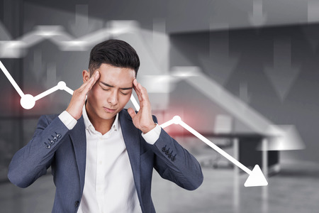 Stressed Asian businessman standing in his office with falling graphs drawn behind him. Concept of...