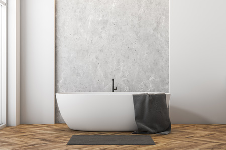 Interior of minimalistic bathroom with white and concrete walls, wooden floor, large window and white bathtub with towel on it and gray rug near it. 3d renderingの写真素材