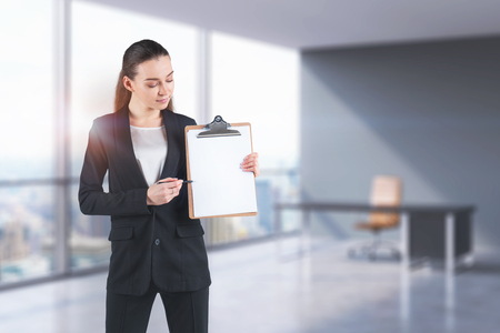 Serious young businesswoman wearing suit showing at her clipboard with pen standing in blurred office. Concept of marketing. Toned image mock upの写真素材