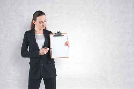 Portrait of young businesswoman with fair hair wearing black suit, holding clipboard and pointing at it with her pen. Concrete wall background. Mock upの写真素材