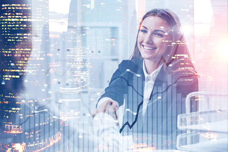 Smiling businesswoman shaking hands in her office over cityscape background with double exposure of graphs. Toned imageの写真素材
