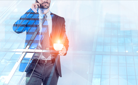 Unrecognizable young businessman talking on smartphone holding cup of coffee over skyscraper background. Toned image double exposureの写真素材