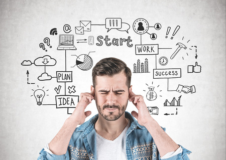 Portrait of bearded young man wearing jeans shirt and thinking hard with eyes closed and fingers on temples standing near concrete wall with start up sketch drawn on itの写真素材
